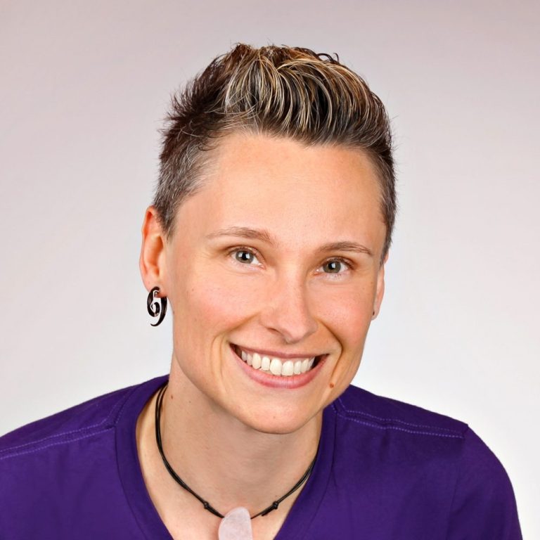 Person mit kurzen, gestuften Haaren und Lächeln, trägt ein lila Shirt und Ohrschmuck.